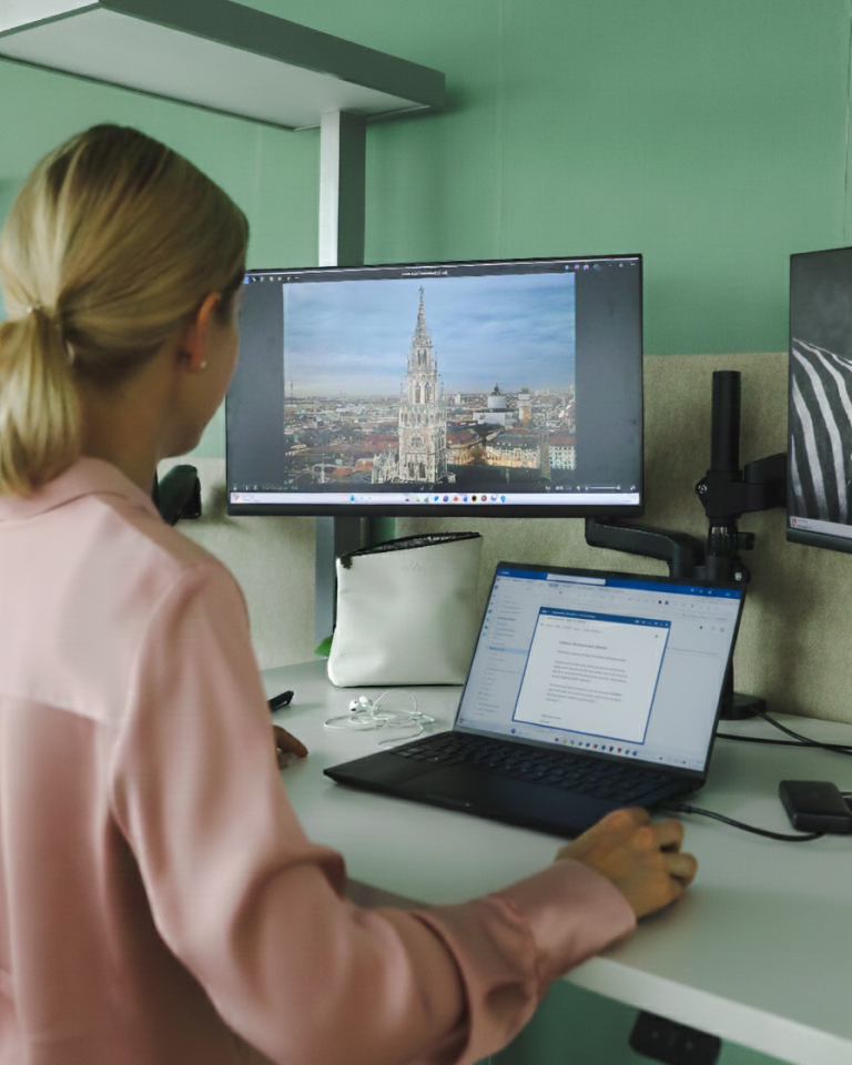 Blonde Frau steht vor zwei Bildschirmen, auf einem ist die Stadt München zu sehen, auf dem anderen ein weißer Bildschirm, elektronische Signaturen